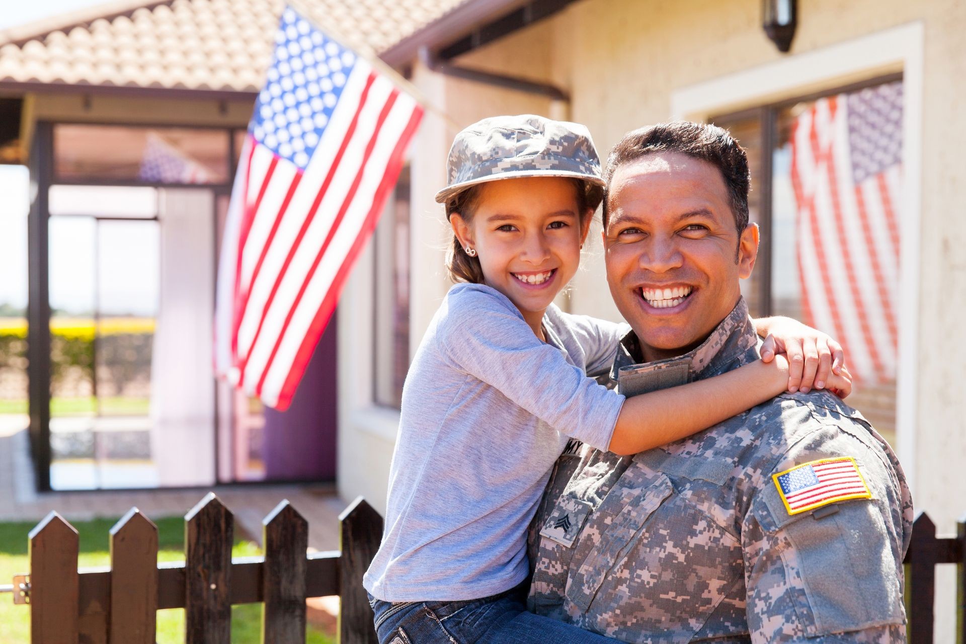 Child embracing person in military uniform, standing in front of house adorned with American flag.